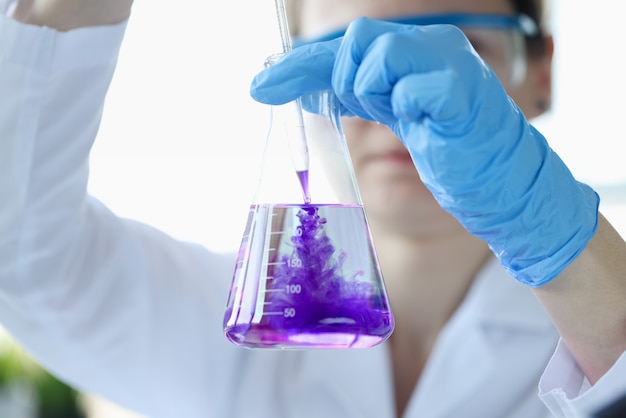 Женщина-ученый капает фиолетовую жидкость в колбу с крупным планом растворителя.