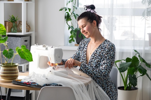 A woman sews tulle on an electric sewing machine in a white modern interior of a house with large windows house plants Comfort in the house a housewife's hobby