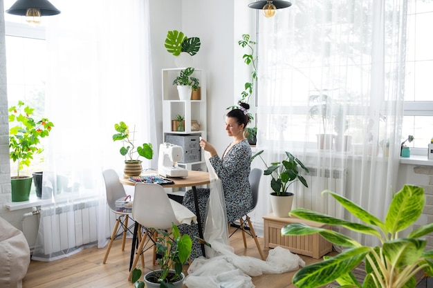 Женщина шьет тюль на электрической швейной машине в белом современном интерьере дома с большими окнами комнатными растениями Комфорт в доме хобби домохозяйки