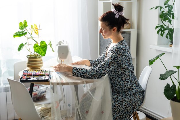 A woman sews tulle on an electric sewing machine in a white modern interior of a house with large windows house plants Comfort in the house a housewife's hobby