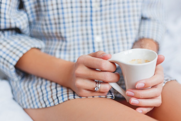 Woman in shirt is sitting on bed with a cup with hot coffee.