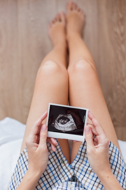 Photo woman in shirt sitting on bed with ultrasound photo of baby in hands. woman is expecting baby. cozy happy morning at home.