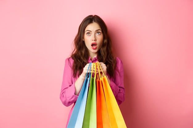 Woman shocked of discounts, looking up with amazed face, holding shopping bags, standing against pink wall.