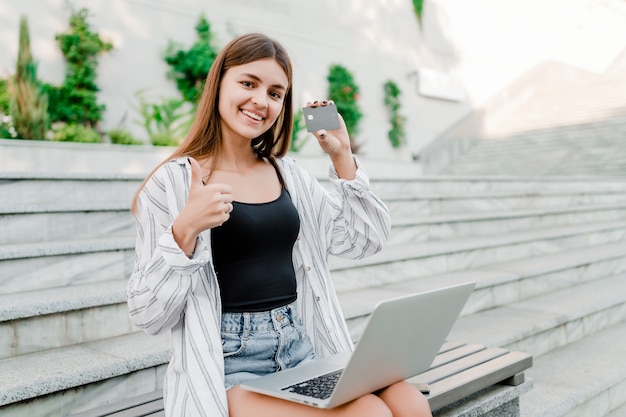 Woman showing credit card and thumbs up with laptop outdoors