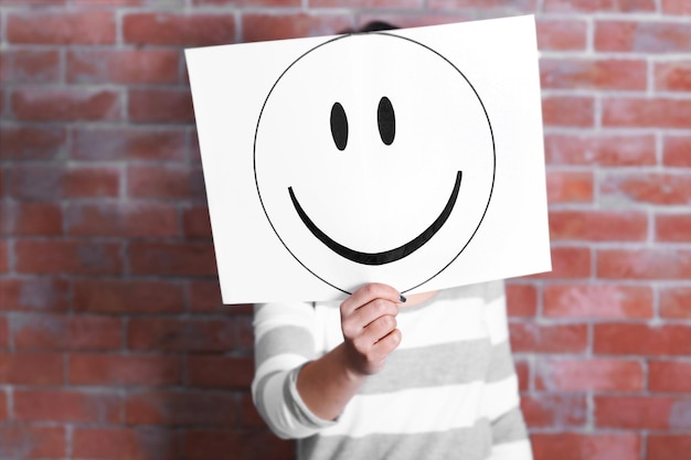 Woman showing a happy emoticon in front of face against a brick wall background