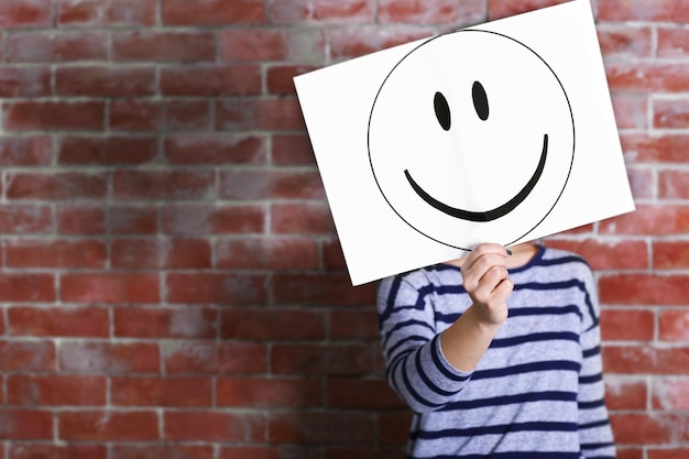 Photo woman showing a happy emoticon in front of face against a brick wall background