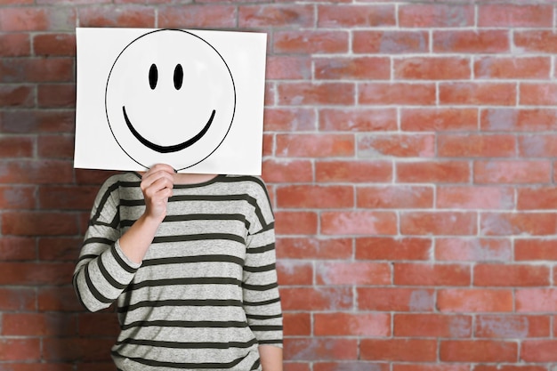 Woman showing a happy emoticon in front of face against a brick wall background