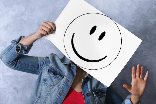 Woman showing a happy emoticon in front of face against a grey textured wall background