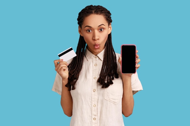 Woman showing mobile phone holding credit bank card online banking looking at camera with shock