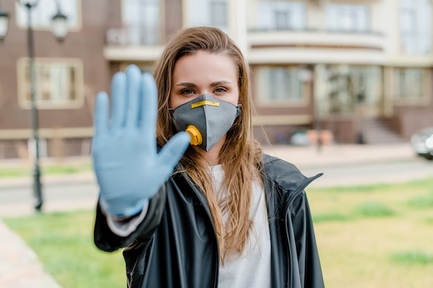 Photo woman showing stop sign with hand wearing respirator mask and gloves outside on the city street