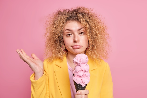 woman shrugs shoulders with clueless expression eats junk food sweet cold ice cream dressed in yellow jacket 