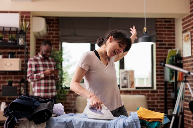 Woman singing loudly effusively while doing house chores,\
african american husband laughs amused in background listening.\
housewife happily doing housework, culturally diverse couple.