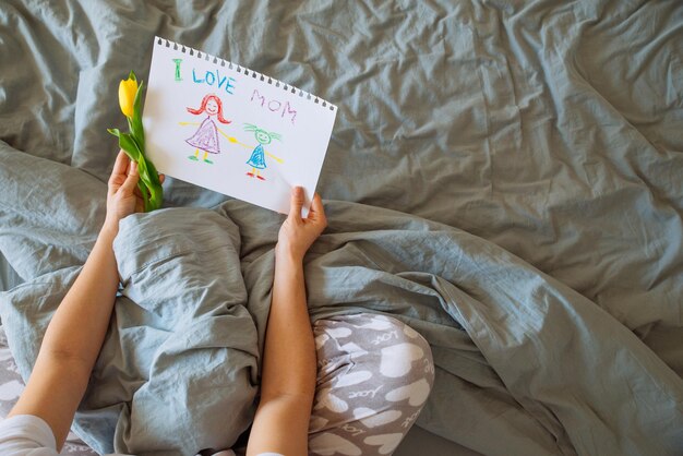 Woman sit in bed and look at picture that kids drew for mother day