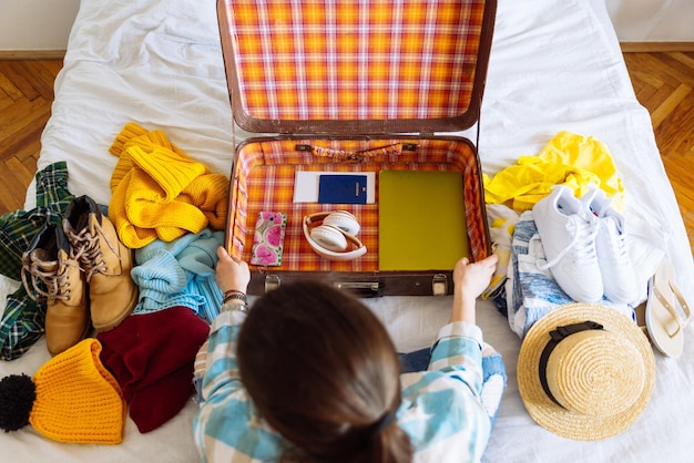 La donna si siede sul letto con valigia e vestiti intorno. concetto di viaggio. decidere il paese caldo o freddo dove andare.