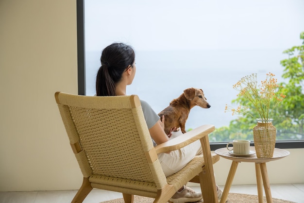 Woman sit on chair with her dachshund dog and enjoy the view outside the window