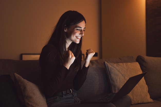 Woman sit at couch read email on laptop makes yes gesture feels happy Female entrepreneur get great business news celebrate career growth advance Achievement win moment of auction victory at nightxA