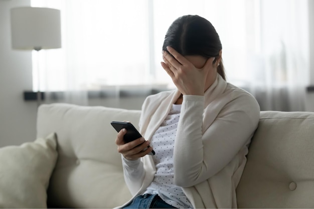 Woman sit on sofa holding smartphone cover face with hand feels scared humiliated suffering from cyberbullying being online abused by stalker Bad news life troubles break up with boyfriend concept