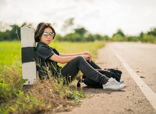 Woman sit with backpack hitchhiking along a road in countryside