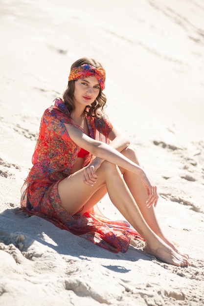 A woman sits on the beach wearing a red dress with a scarf on it.