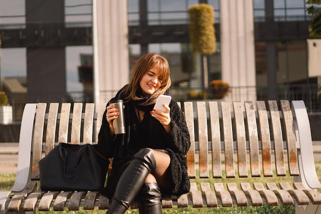 街のベンチに座っている女性がサーモカップからお茶を飲み、電話を使って