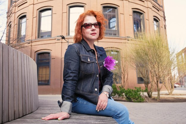 A woman sits on a bench in front of a building with a flower on her jacket.