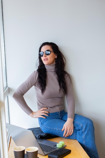A woman sits on a bench in front of a window wearing a turtleneck and jeans.