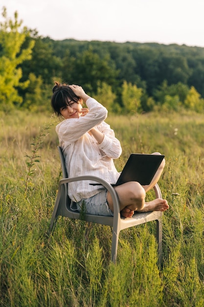 A woman sits on a chair in a field with a laptop on her lap.