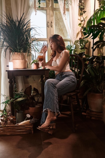 A woman sits in a chair in front of a window with plants and a potted plant.