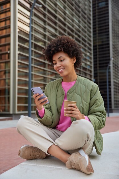 woman sits crossed legs takes rest holds mobile phone checks email box browses websites drinks takeaway coffee dressed in stylish clothes