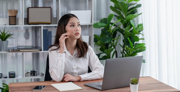 A woman sits at a desk in an office, looking at a laptop and a plant behind her.