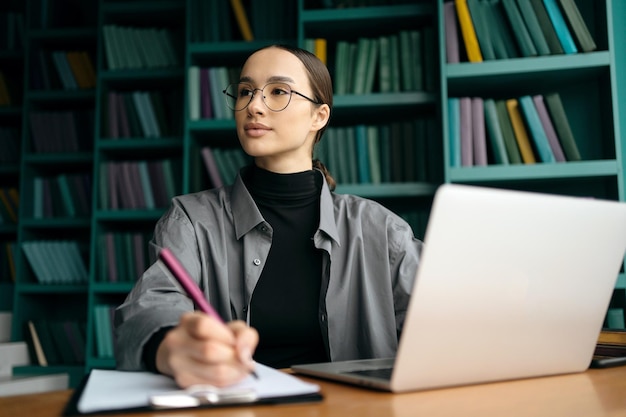 A woman sits at a desk with a laptop and a bookcase in the background.