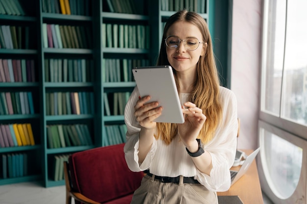 A woman sits in front of a bookshelf and reads a tablet.