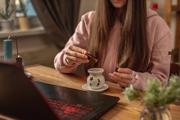 Woman sits in front of a laptop listens to an instructor or a video lecture on meditation and relaxation and drips essential oil into an aroma lamp Concept of a remote therapy and meditation session