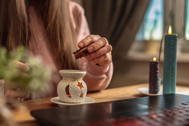 Woman sits in front of a laptop listens to an instructor or a\
video lecture on meditation and relaxation and drips essential oil\
into an aroma lamp concept of a remote therapy and meditation\
session