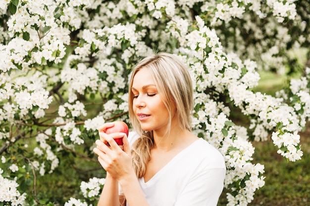 Foto una donna si siede sull'erba sotto gli alberi in fiore e tiene in mano una mela rossa, lunghi capelli biondi, bellezza