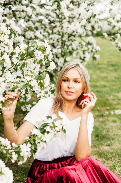 Una donna si siede sull'erba sotto gli alberi in fiore e tiene in mano una mela rossa, lunghi capelli biondi, bellezza