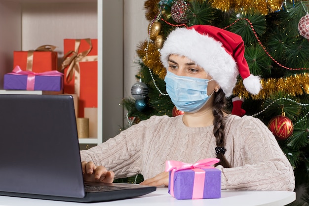 A woman sits near a laptop in a Santa Claus hat and mask.