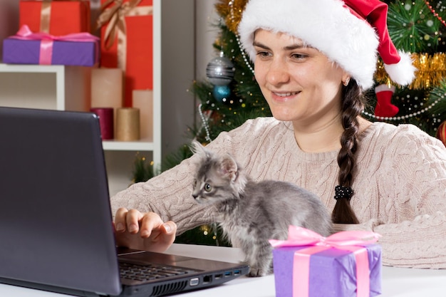 A woman sits near a laptop in a Santa Claus hat.