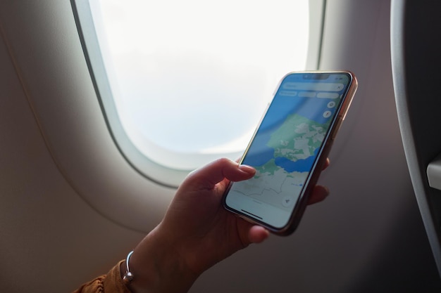 Woman sits near the porthole in an airplane and looks at a map on a phone Travel and smartphone concept Girl holds smart phone