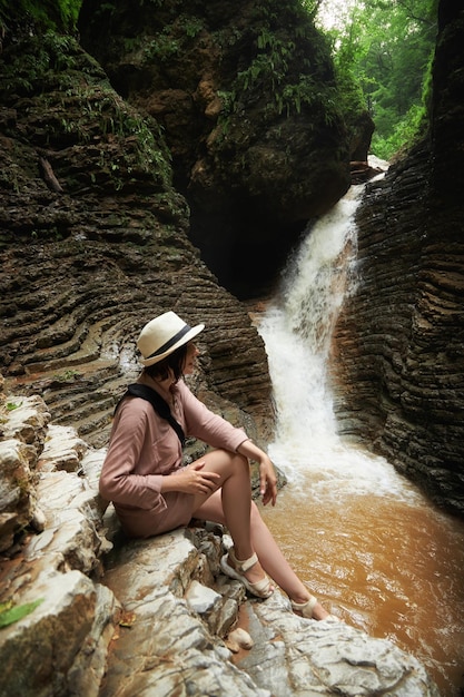 A woman sits on a rock and looks at a waterfall in the forest a woman in a hat a vacation in nature
