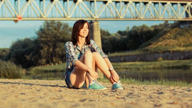 a woman sits in the sand in front of a bridge