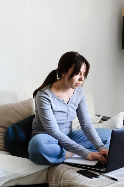 A woman sits on a sofa, works on a laptop, types on a keyboard\
and writes notes in a notebook.