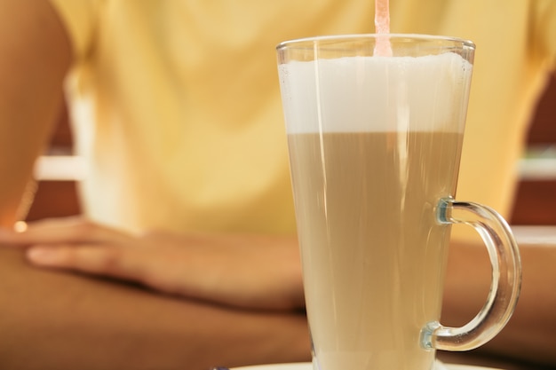 Woman sits at a table in a cafe and drinking latte in a glass with straw closeup