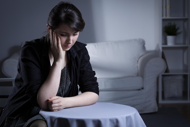 A woman sits at a table in front of a white couch and looks sad.