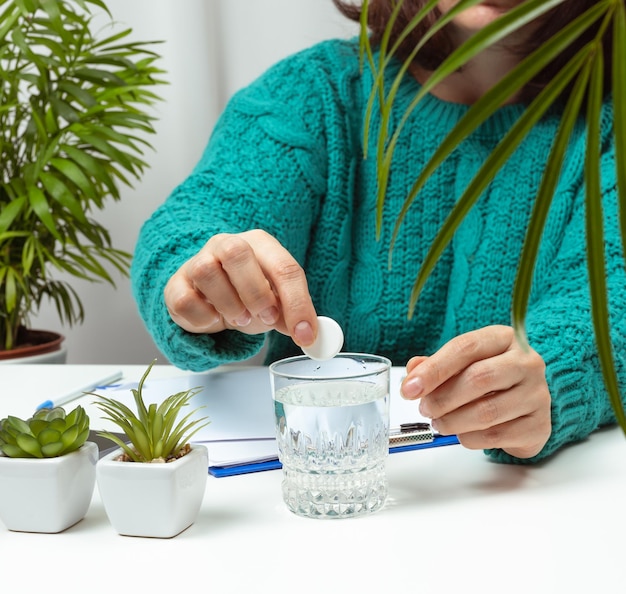 Woman sits at the table and throws a round aspirin into the sakan with water, headache