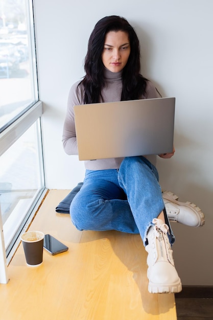 A woman sits on a table with a laptop and looks at a coffee cup.