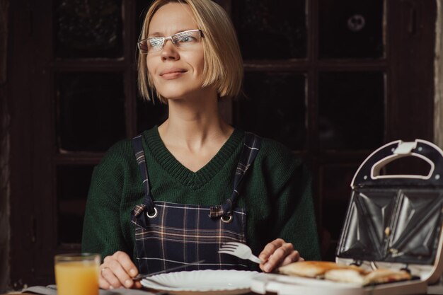 女性はサンドイッチ メーカーと皿を持ってテーブルに座ります 朝食の軽食 中年女性 適切な栄養