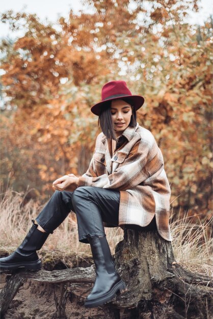 Foto una donna siede su un tronco d'albero con un cappello rosso e un cappotto scozzese.