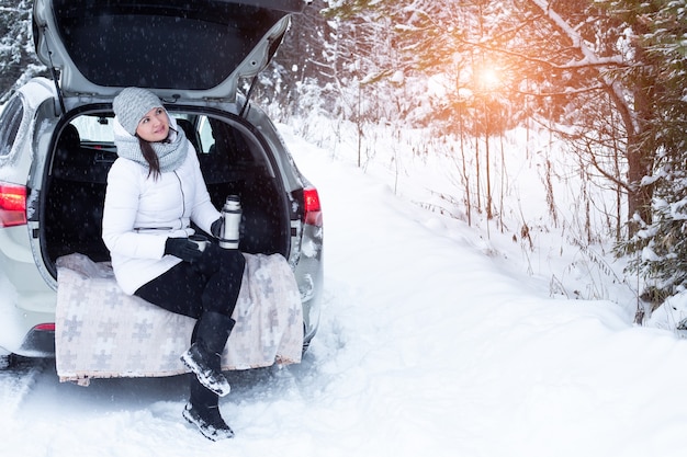 Woman sits in trunk of the car and holds a cup of hot tea in hands. Winter vacation, travel. Snow forest and road. Snowy weather - Image