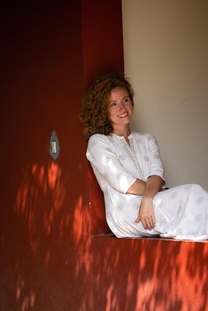 A woman sits on a wall in a white dress and smiles.